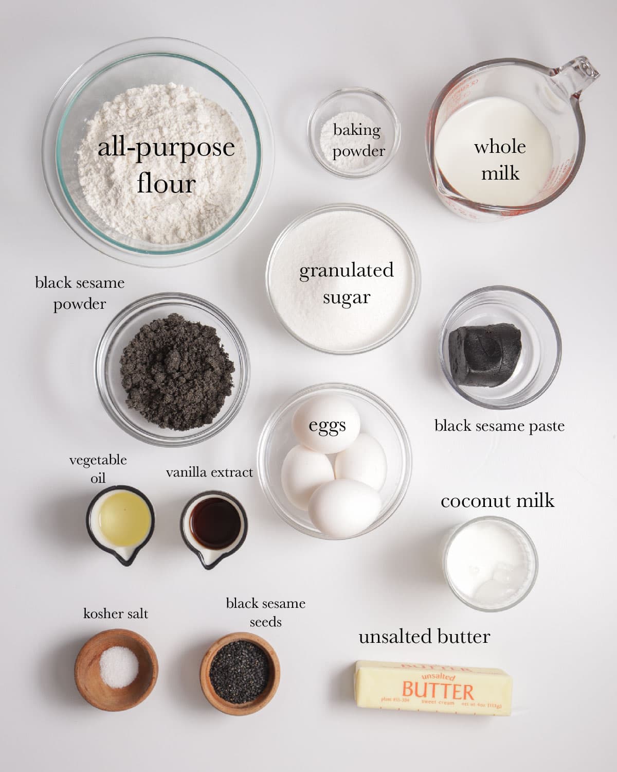 overhead view of ingredients top to bottom: all-purpose flour, baking powder, whole milk, black sesame powder, granulated sugar, black sesame paste, vegetable oil, vanilla extract, eggs, coconut milk, kosher salt, black sesame seeds, unsalted butter