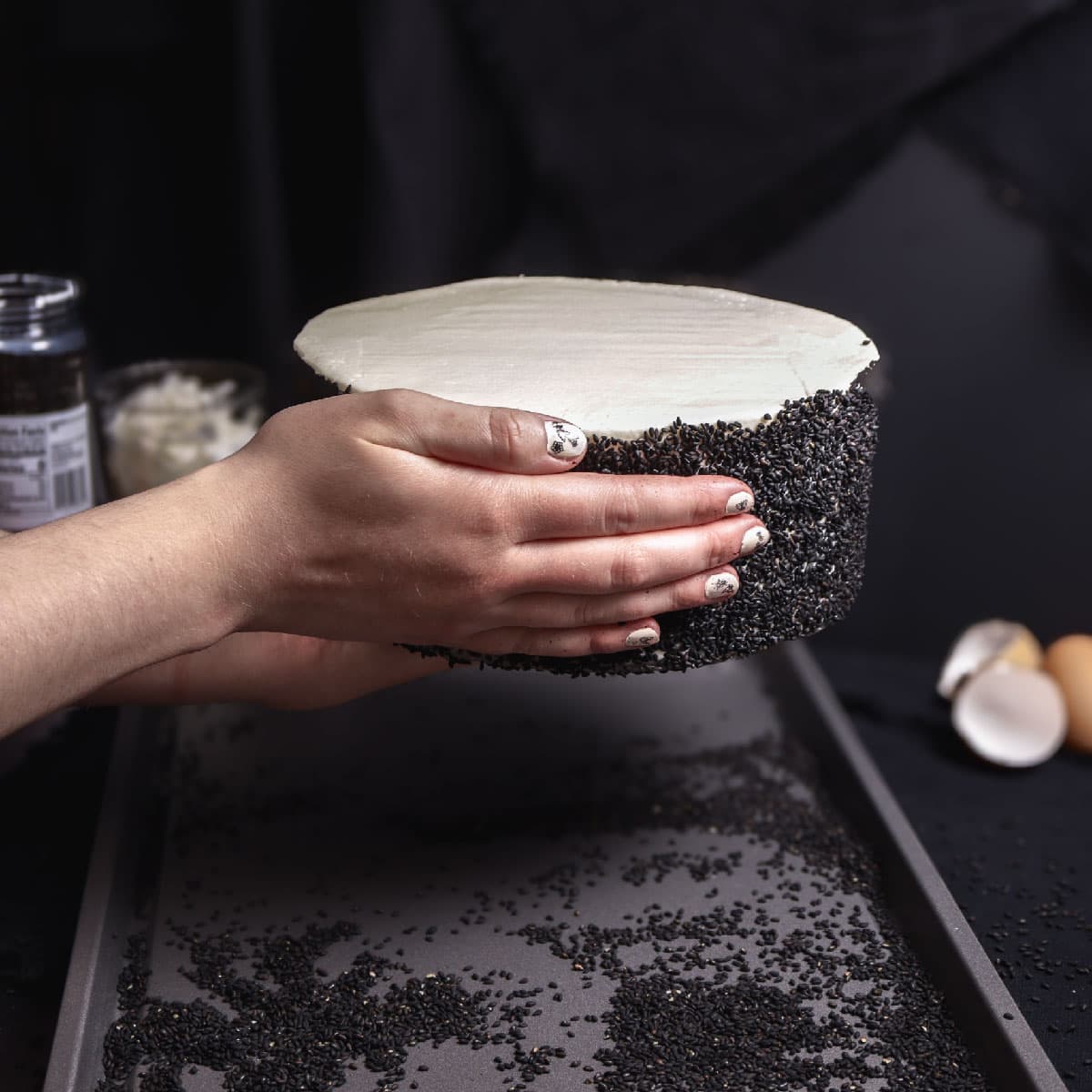 pushing black sesame seeds into the edge of a round layer cake with white frosting