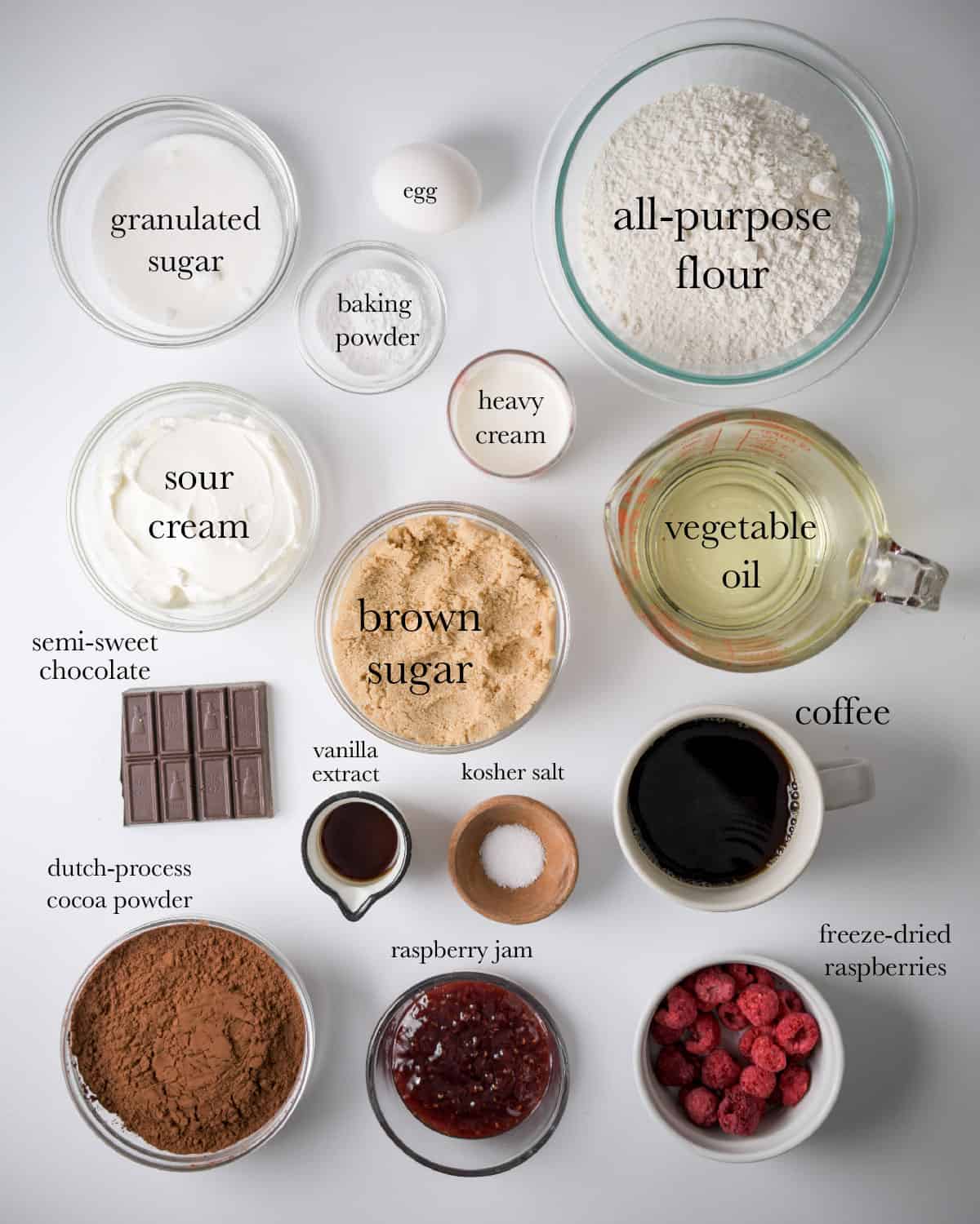 ingredients overhead view top to bottom: granulated sugar, egg, all-purpose flour, baking powder, heavy cream, sour cream, brown sugar, vegetable oil, semi-sweet chocolate, vanilla extract, kosher salt, coffee, dutch-process cocoa powder, raspberry jam, freeze-dried raspberries