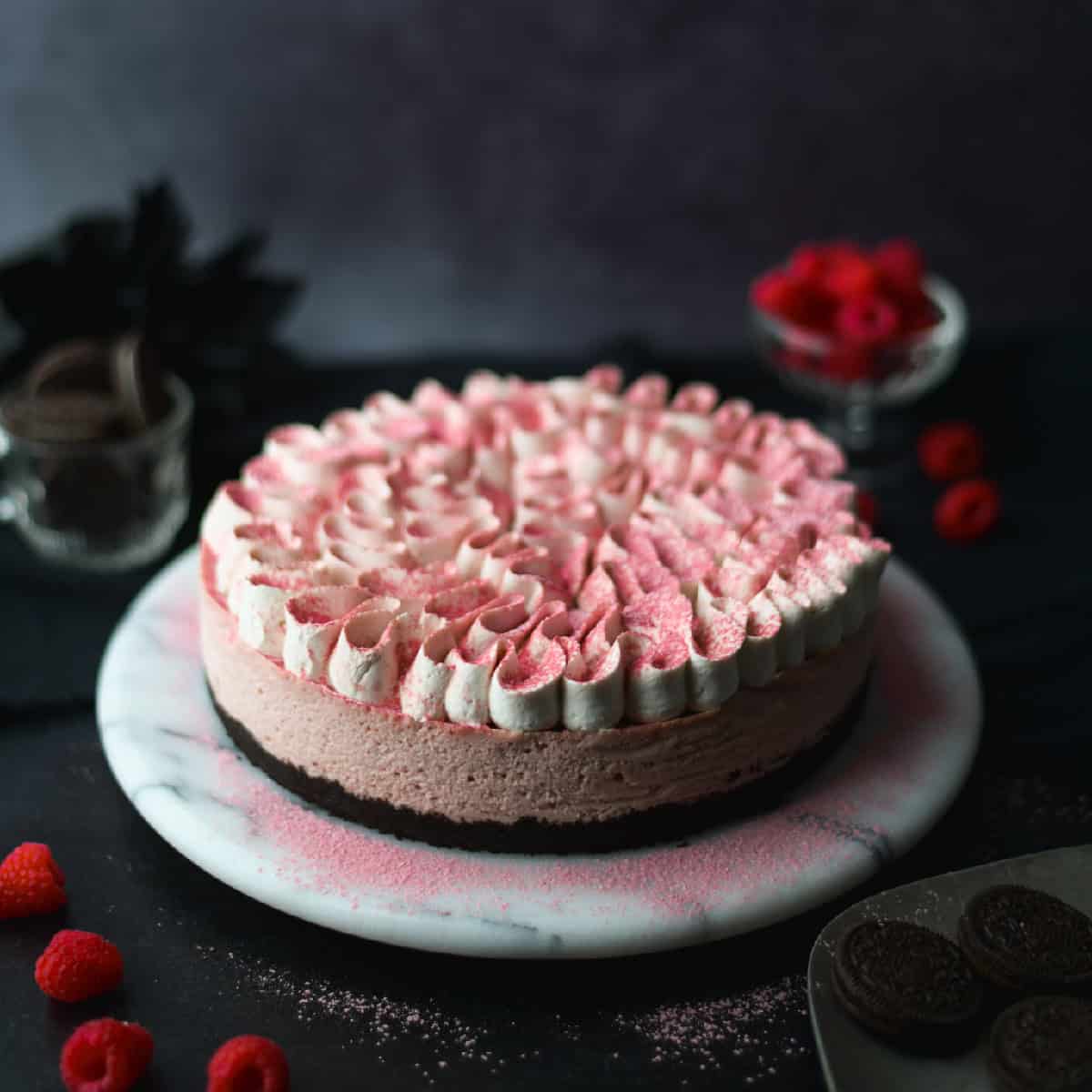 raspberry cheesecake on white marble turntable, black background, raspberries and oreos nearby
