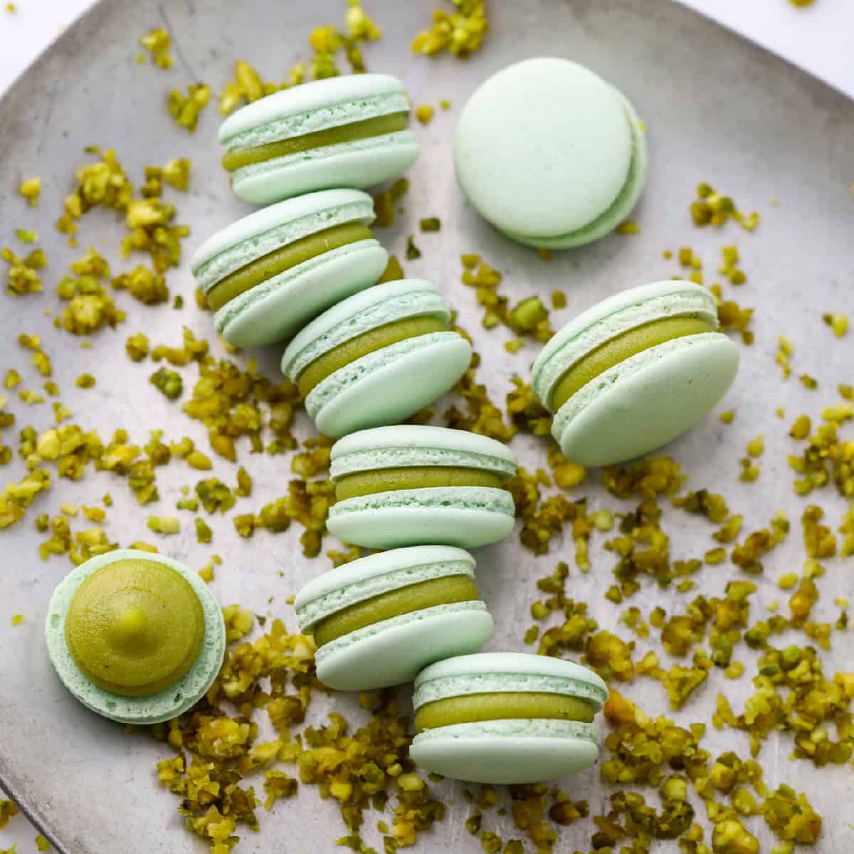 pistachio macarons with pistachio filling on a silver tray with ground pistachios nearby.