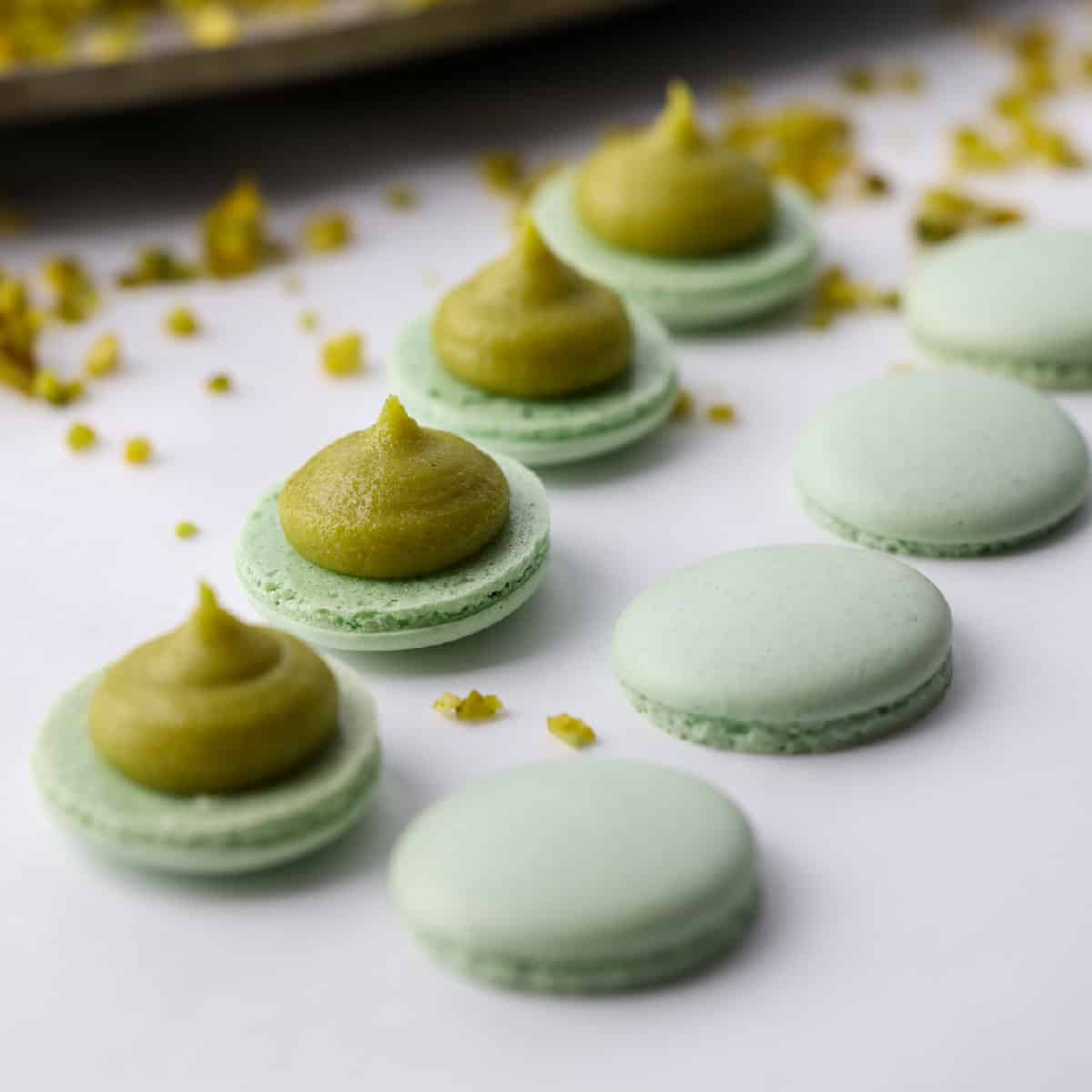 pistachio macarons in a line with pistachio filling piped on the bottoms, top shells nearby.